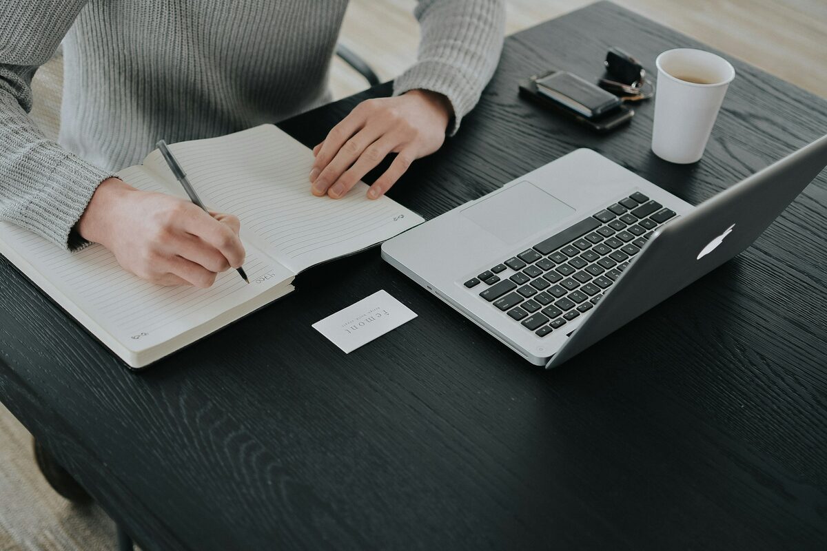A person writing in a notebook beside a laptop at a desk